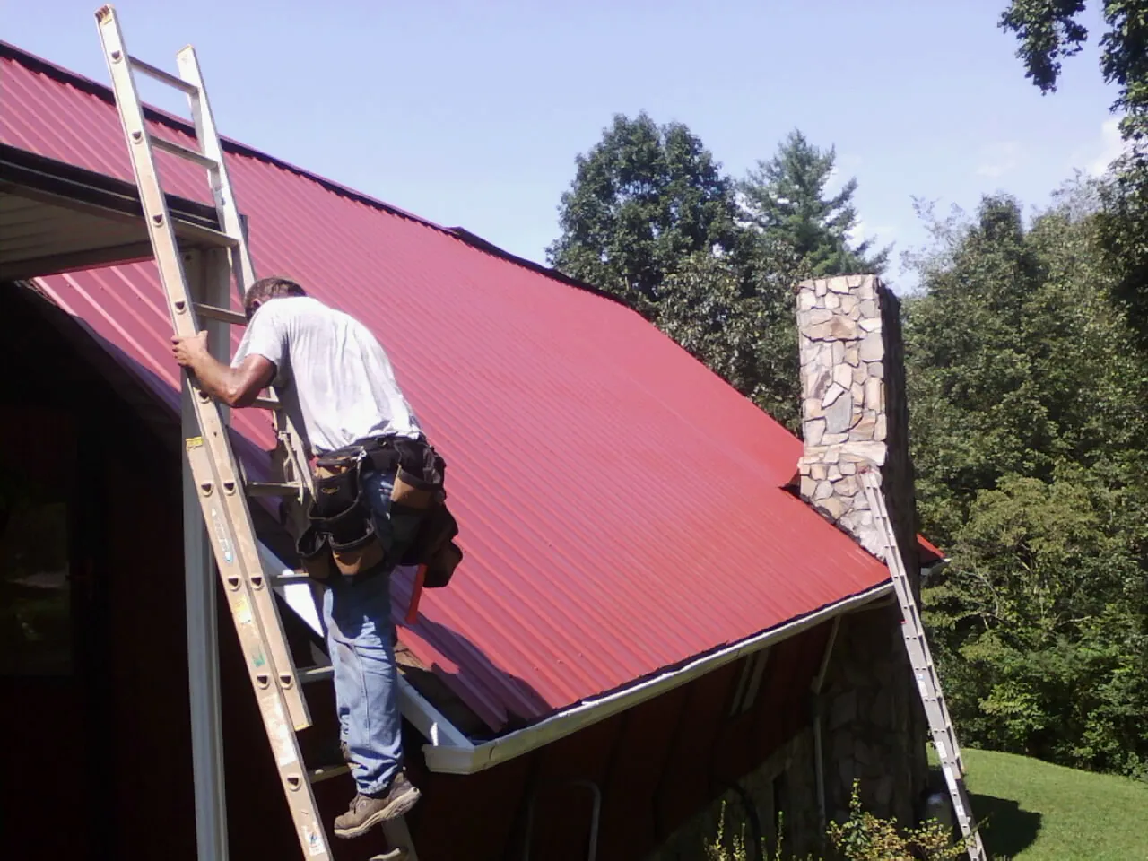 Roofer on a ladder beside a red metal roof and stone chimney — Soffit & Fascia Repair by Roofing Brothers in Vassar